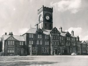 A Ford Popular parked outside as a chap stood with his bike lights a cigarette. The front doors are open, welcome to High royds hospital on this fine Summers day in June 1965.